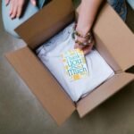 A person packing a white t-shirt with a thank you card into a cardboard box.
